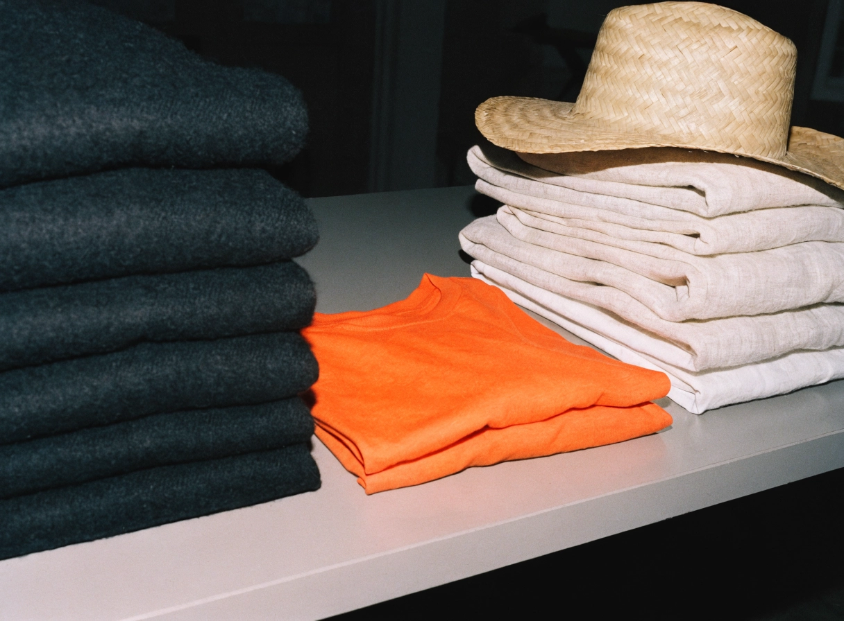 Stack of winter knitwear beside folded summer linen and a straw hat, an orange tee between them