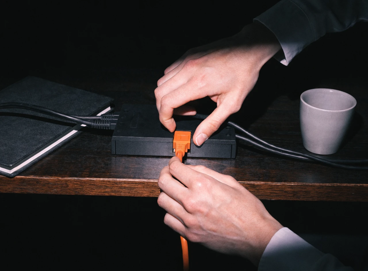 Hands plugging an orange network cable into a black switch on a desk
