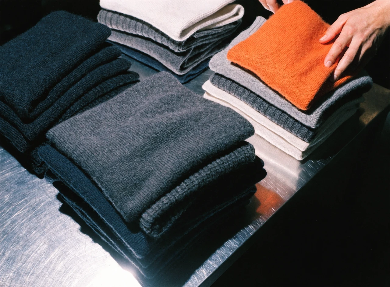 Hands sorting folded knitwear on a steel table, one orange sweater among grey ones